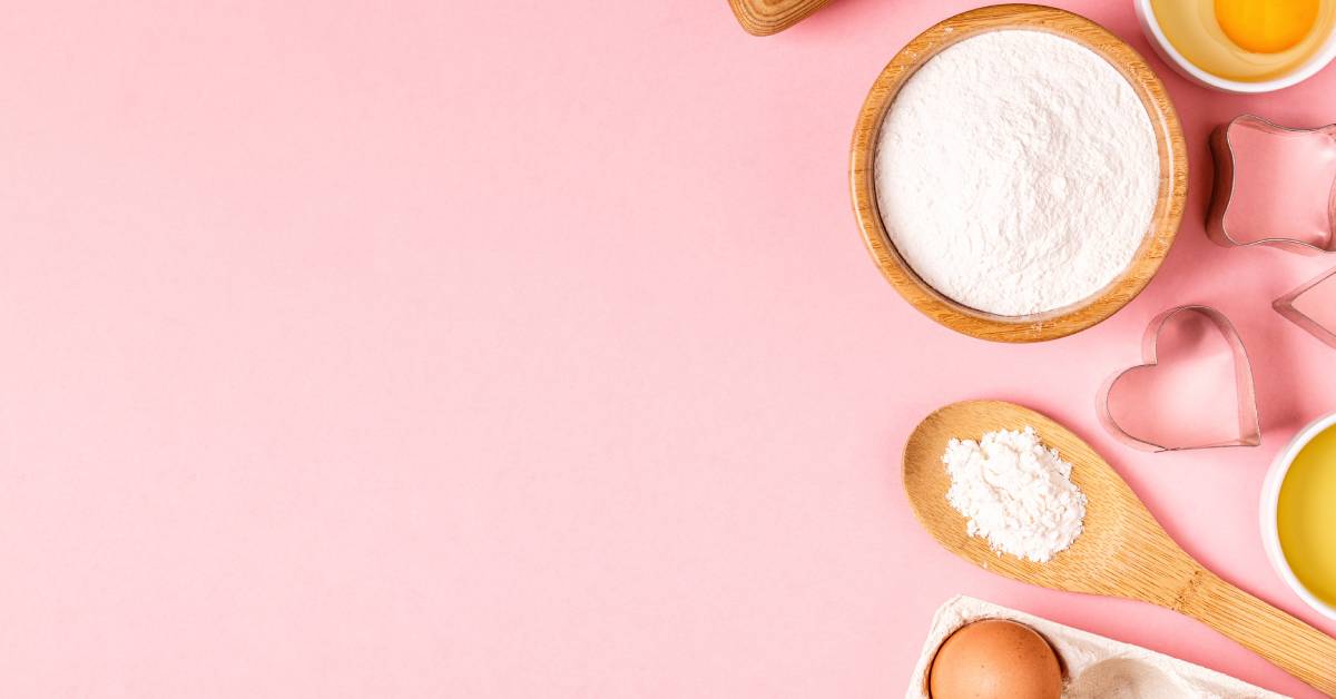 Wooden baking tools and ingredients, including eggs and flour, arranged on a pastel pink background.