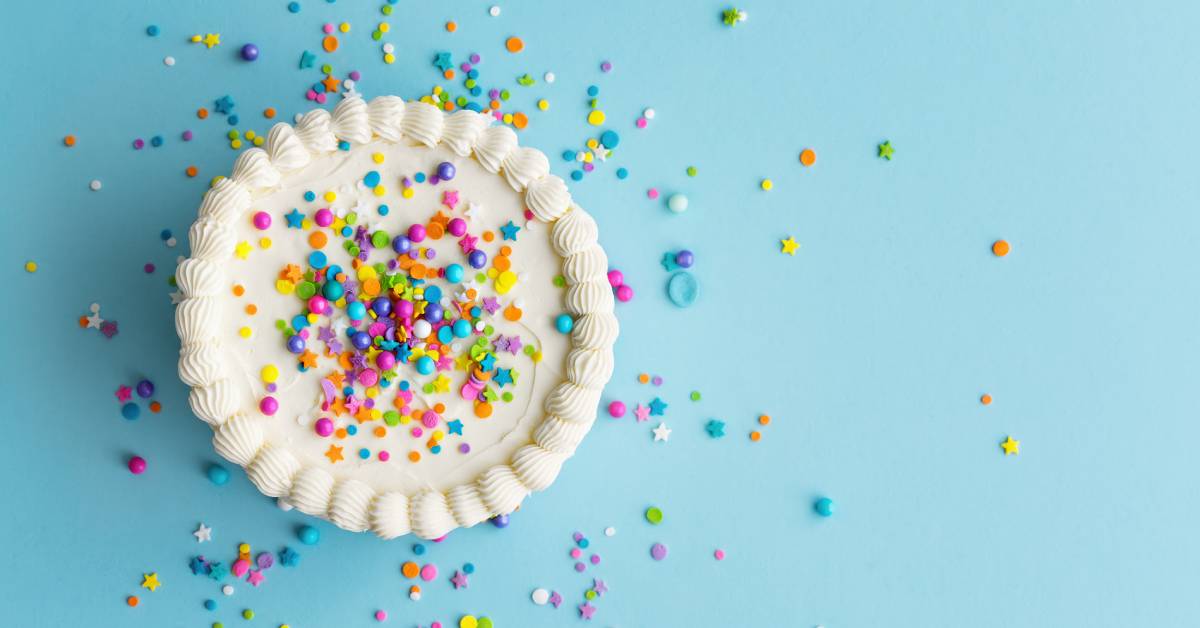 A white frosted cake decorated in vibrant round and star-shaped sprinkles. The cake is on a light blue surface.