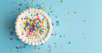 A white frosted cake decorated in vibrant round and star-shaped sprinkles. The cake is on a light blue surface.