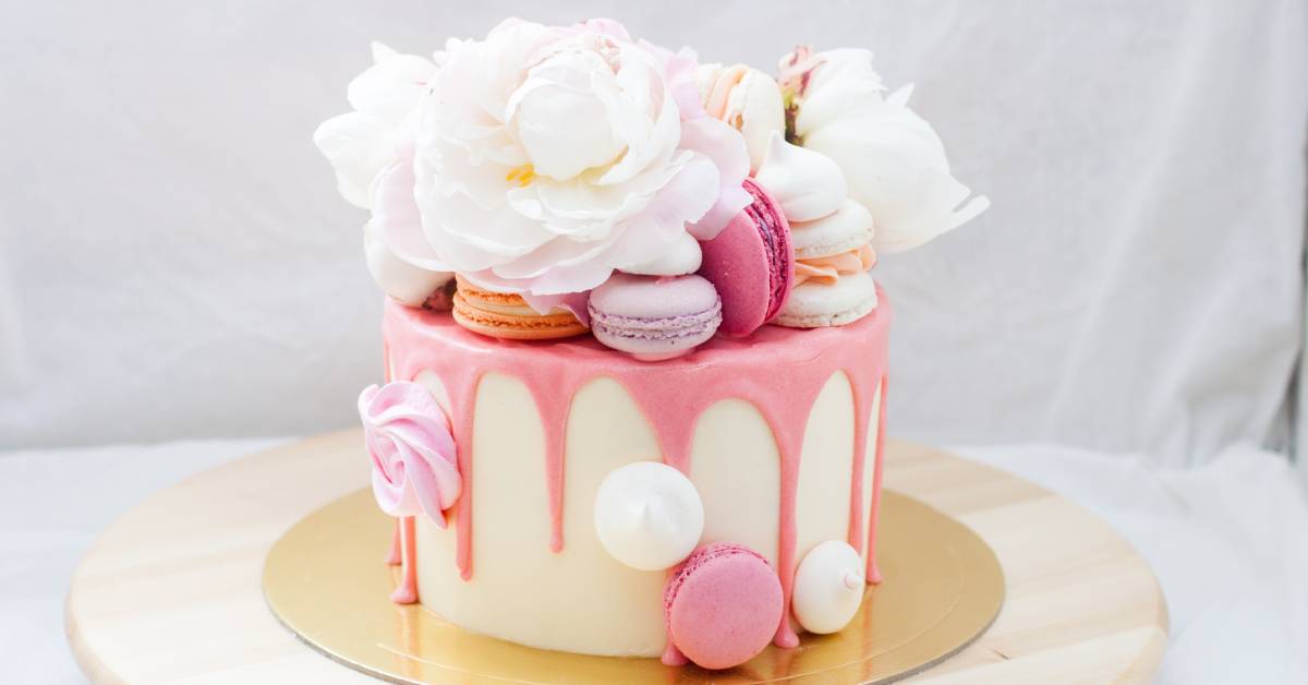  A white cake on a gold plate topped with melted pink chocolate, pink and white macaroons, and pink and white flowers.