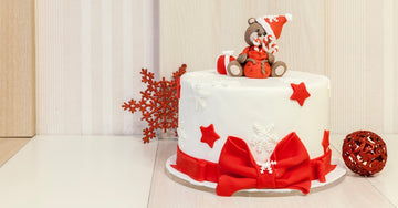 A round, white cake decorated with a red fondant ribbon, stars, snowflakes, and a teddy bear in a Santa hat.