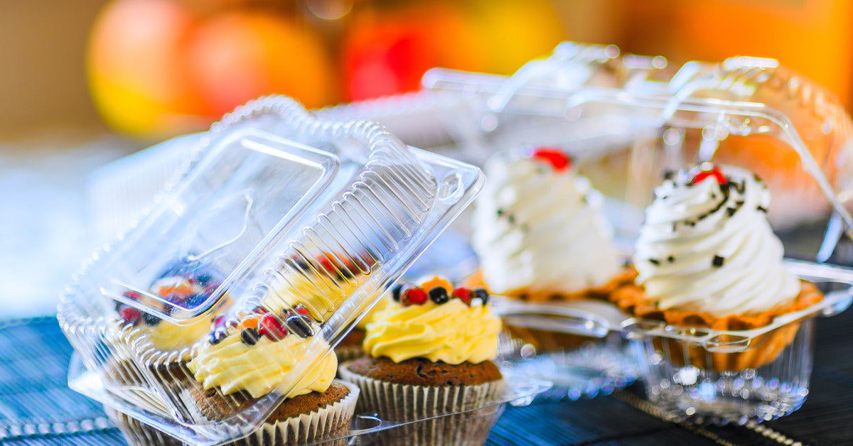 Plastic cupcake containers sitting open on a table. The cupcakes are decorated with white frosting, fruit, and more.