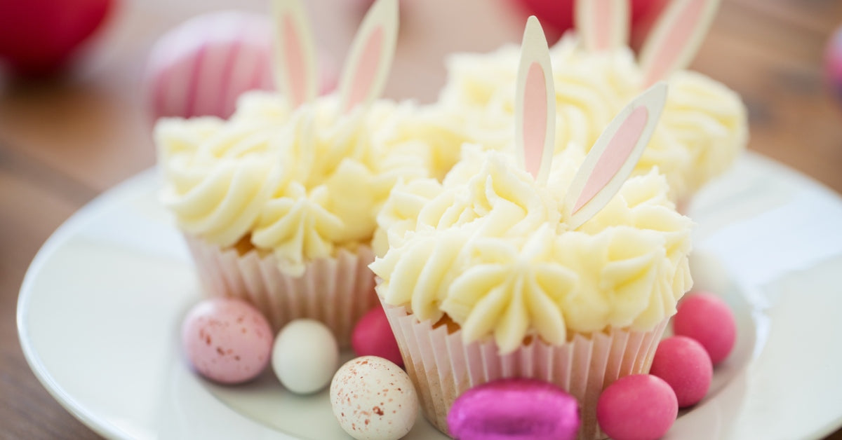 A plate of cupcakes decorated with white frosting and bunny ears. There are pink and white Easter eggs on the plate, too.