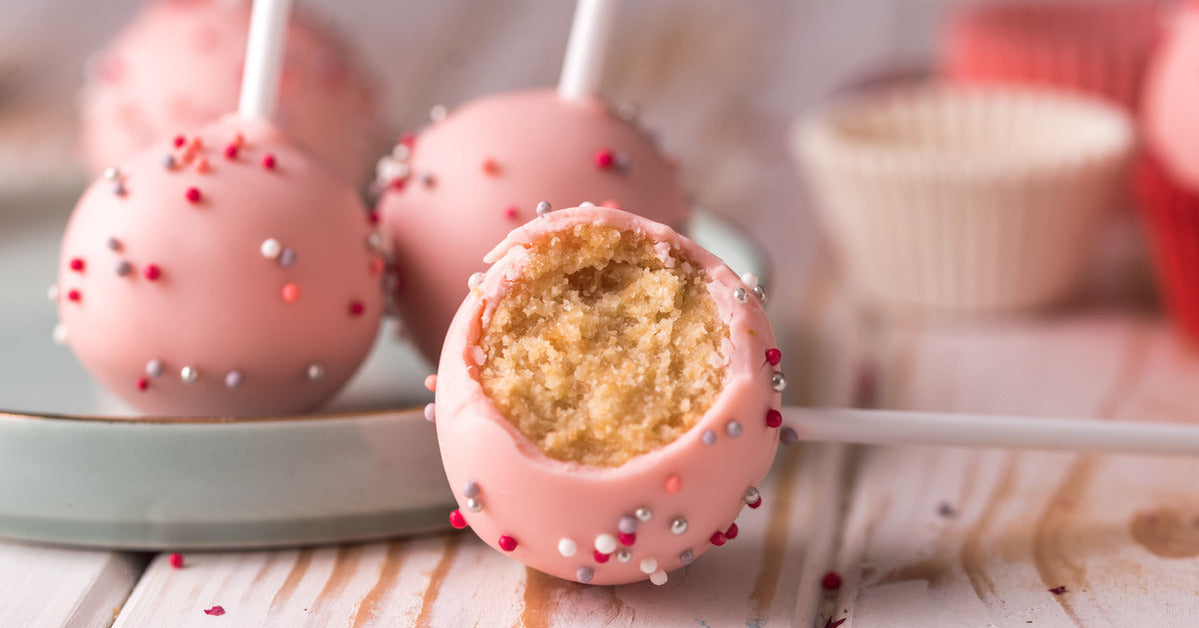 Cake pops with pink coating and pink, red, silver, and white sprinkles. One is laying down with a bite taken out of it.