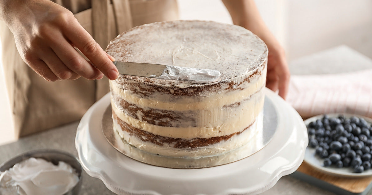 A cake decorator applies a white crumb coat on a cake using a thin spatula. There is frosting and blueberries on the counter.