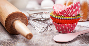 Baking tools and utensils on a flour-covered table. There is a rolling pin, a whisk, a spoon, and a stack of cupcake liners.