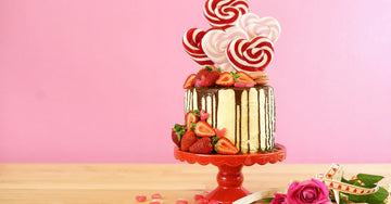 A cake decorated with white frosting, a dripping chocolate glaze, fresh sliced strawberries, and heart-shaped lollipops.