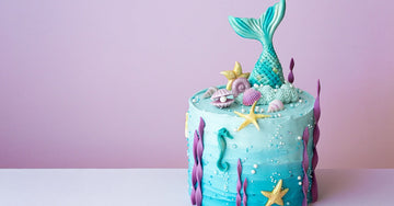 A mermaid-themed cake with ocean-colored frosting, starfish, shells, pearls, and a mermaid tail sticking out of the top.